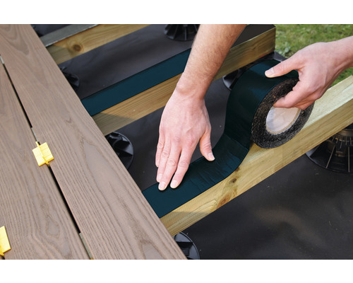 Een persoon bevestigt een afdichtband op een houten onderconstructie voor een terras.