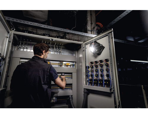 Een man werkt aan een open schakelkast met een werklamp