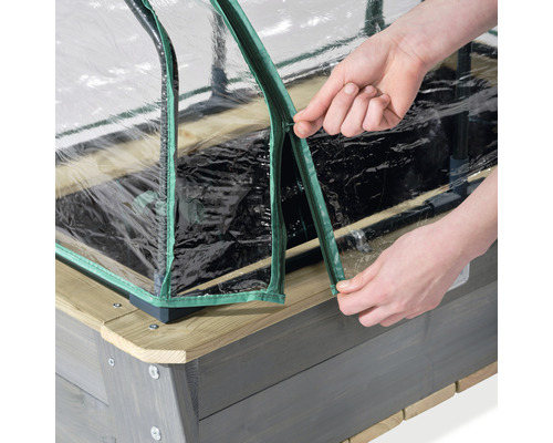 Detailopname van een verhoogde tuinbak met doorzichtige afdekking en ritssluiting, die door handen wordt geopend.