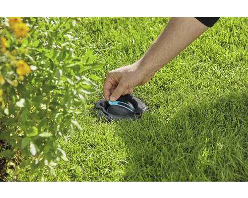 Een persoon installeert een verzonken gazonsproeier in een gazon vlakbij een bloeiende struik.