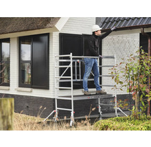 Een persoon staat op een steiger en werkt aan een huis.