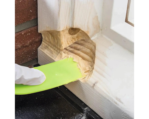 Een hand met handschoen gebruikt een plamuurmes om houtplamuur aan te brengen.