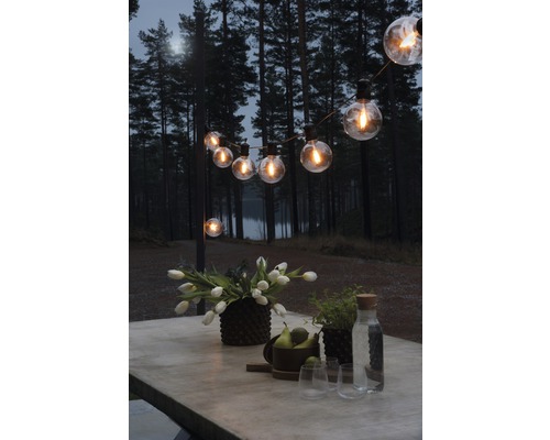 KONSTSMIDE Kerstverlichting LED lichtsnoer retro 10 lampjes warmwit 4,5 m Verlichte buitenruimte met lichtsnoer, tafel, bloemen en drankjes tegen een bosachtergrond