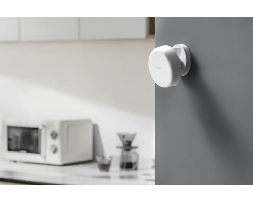 Een witte Aqara deur- en raamsensor is op een grijze muur gemonteerd. Op de achtergrond staat een magnetron.