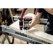 Een persoon gebruikt een Metabo bovenfrees op een Metabo werkbank, aangesloten op een stofzuiger.