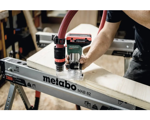 Een persoon gebruikt een Metabo bovenfrees op een Metabo werkbank, aangesloten op een stofzuiger.