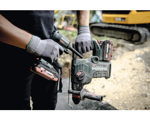 Een man houdt een Metabo combihamer en een patroonpistool in zijn hand, op de achtergrond is een graafmachine te zien