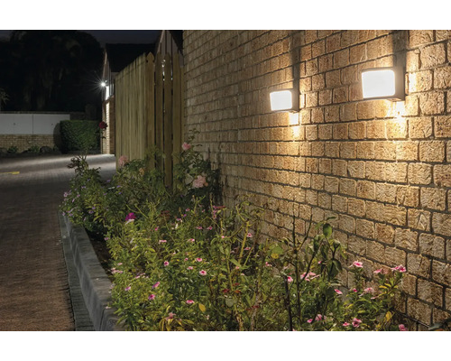 Twee vierkante buitenlampen aan een bakstenen muur naast beplanting.