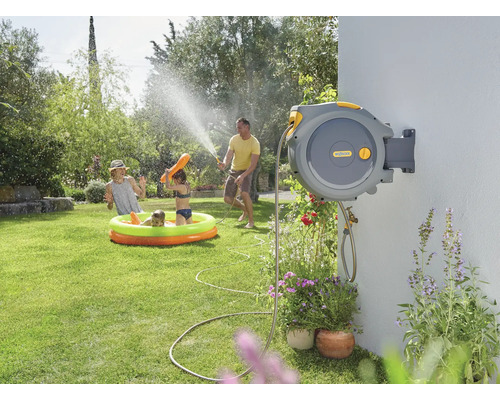 Tuinscene met oprolbare slangbox aan een muur, spelende kinderen in een opblaasbaar zwembad en een volwassene die ze besproeit met een tuinslang.