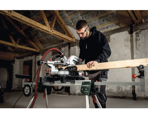 Man met veiligheidsbril zaagt hout met een Metabo verstekzaag op een onderstel in een ruwbouw.