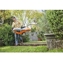 Een vrouw draagt een grasmaaier door een tuin.