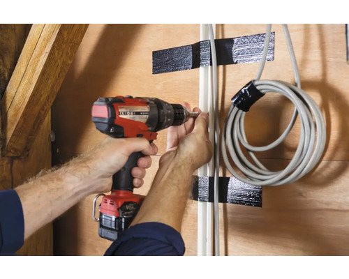 Een persoon bevestigt met een accuschroevendraaier een kabelgoot aan een houten wand, waarbij een opgerolde kabel eveneens aan de wand is bevestigd.