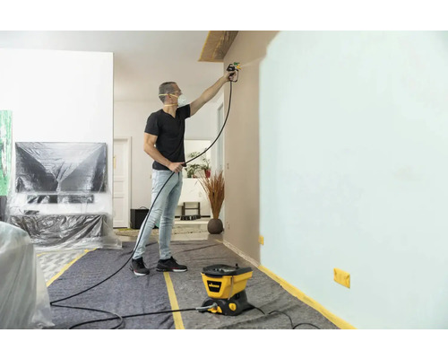 Man schildert muur met verfspuitsysteem in een woonkamer beschermd met afdekmateriaal.