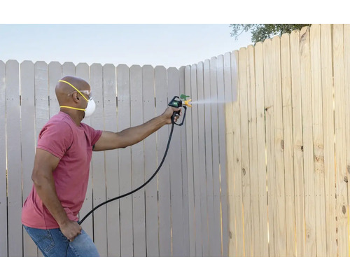 Iemand schildert een houten schutting met een verfspuit, waarbij hij een veiligheidsmasker en een veiligheidsbril draagt.