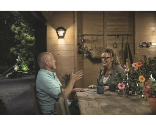 Een echtpaar zit aan een houten tafel in een helder verlicht tuinhuis met een buitenwandlamp.