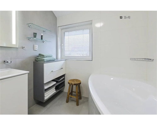 Lichte badkamer met ligbad, wastafel, onderkast, handdoeken, houten kruk en raam met jaloezie