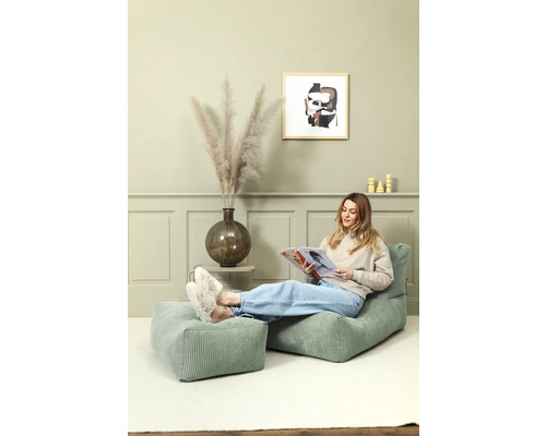 Vrouw leest op een groene corduroy stoel met voetenbank in een lichte kamer.