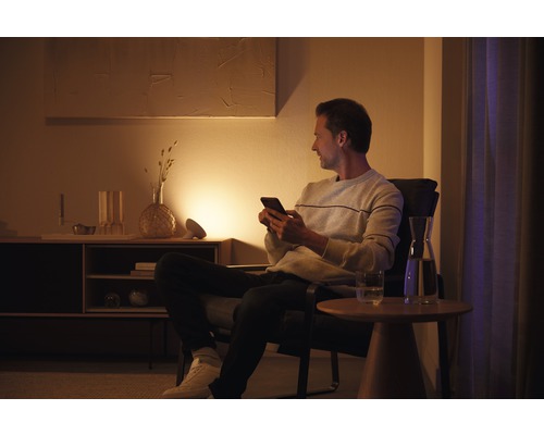 Een man zit met een smartphone in een fauteuil in een woonkamer met dressoir, lamp en tafel.