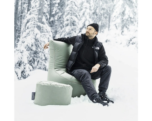 Man zit in een lichte stoel met voetenbank in de sneeuw voor bomen