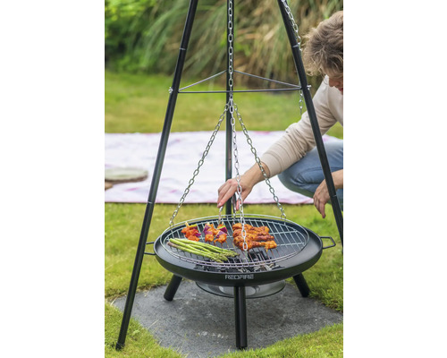 Persoon grilt op een driepootbarbecue met een grillrooster waaraan een ketting is bevestigd. Op het rooster liggen spiesjes, vlees en groene asperges.