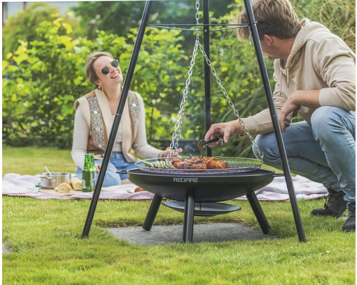 Scene met een Redfire driepoot barbecue in de tuin met twee personen die eten aan het grillen zijn.