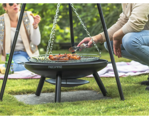Drie personen barbecueën op een driepootbarbecue in de tuin met vlees en groenten.