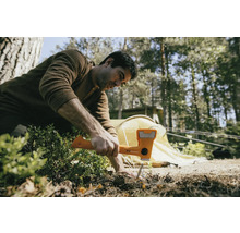 Een man gebruikt een hamer om een haring in de grond te slaan, met een tent op de achtergrond.