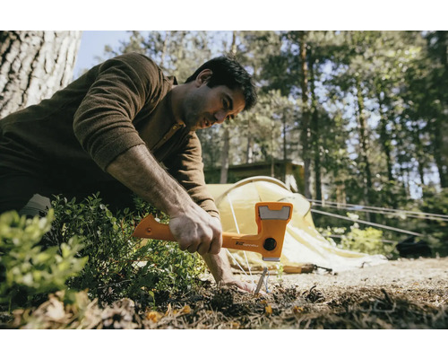 Een man gebruikt een hamer om een haring in de grond te slaan, met een tent op de achtergrond.
