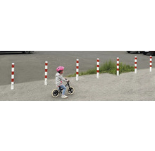 Meerdere rood-wit gestreepte verkeerspalen begrenzen een gebied, terwijl een kind er met een loopfiets voorbij rijdt.