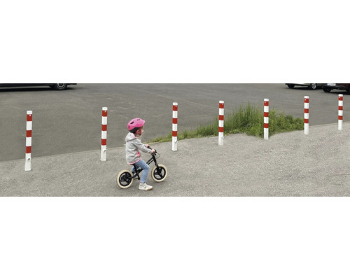 Meerdere rood-wit gestreepte verkeerspalen begrenzen een gebied, terwijl een kind er met een loopfiets voorbij rijdt.
