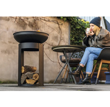 Metalen vuurschaal met houtvak in de tuin en vrouw die een drankje drinkt