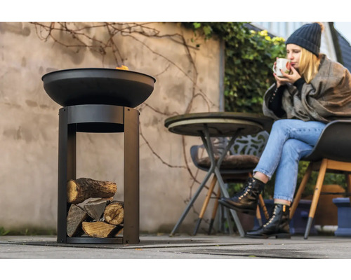 Metalen vuurschaal met houtvak in de tuin en vrouw die een drankje drinkt