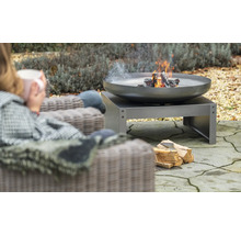 Vuurschaal met hout op een terras, ernaast een persoon in een stoel