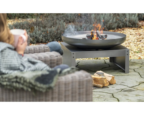 Vuurschaal met hout op een terras, ernaast een persoon in een stoel
