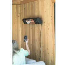 Een infraroodstraler is aan de houten wand gemonteerd, een persoon met een mobiele telefoon is in beeld.