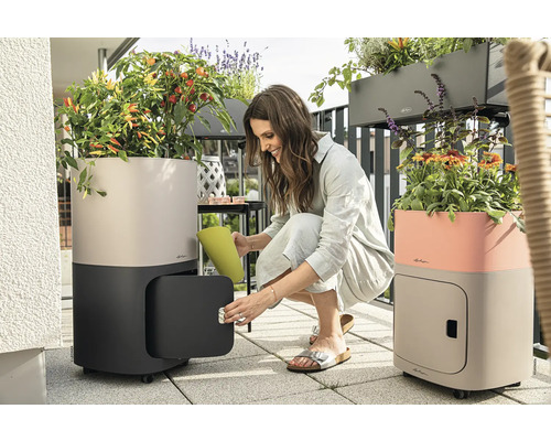 Scene op een balkon met twee plantenbakken met bewateringssysteem, op wielen en met een kastcompartiment, een vrouw geeft planten water