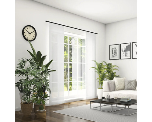 Lichte woonkamer met bank, salontafel, kamerplanten, klok en schuifgordijn voor het raam.