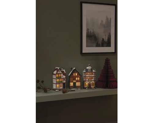 Decoratieve miniatuurhuisjes en een papieren kerstboom op een plank met een ingelijste foto aan de muur