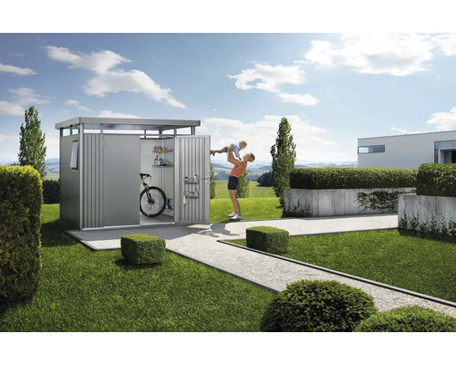 Tuinscene met gereedschapsschuur, fiets, planken en een vrouw die een kind omhoog houdt