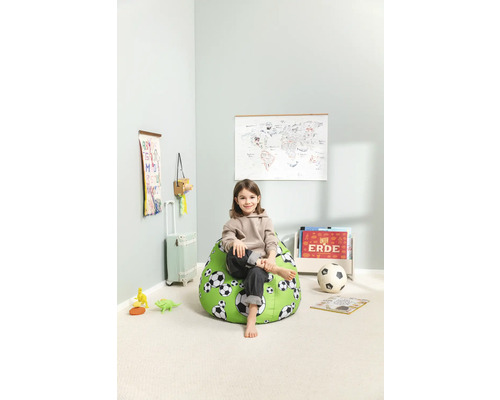 Een kind zit op een groene zitzak met voetbalmotief in een lichte kamer.