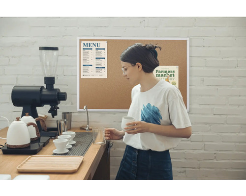 In een lichte cafésetting staat een vrouw met een bril achter de bar, die een kopje en een maatbeker vasthoudt, omringd door een koffiemachine, waterkokers en een menubord aan een bakstenen muur.