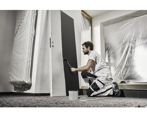 Een man schildert een deur met een verfroller in een kamer die is afgedekt met beschermende folie. Schilderwerkzaamheden