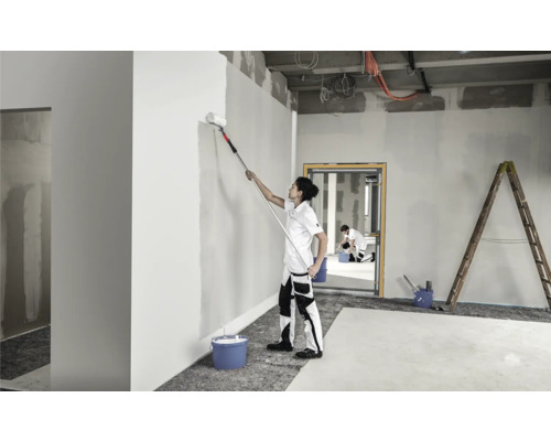 Een persoon schildert een muur in een kamer met een verfroller op een telescoopsteel. Op de achtergrond zijn nog een persoon en een ladder te zien.