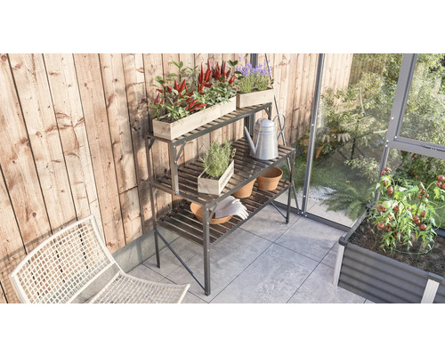 Metalen plantentafel met drie niveaus met planten, potten, gieter en tuinhandschoenen op een balkon naast een kas.