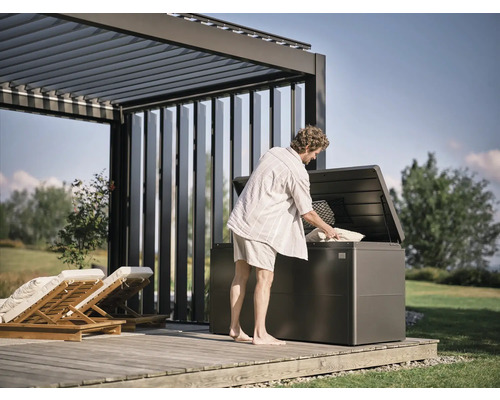 Man vult een tuinkist onder een pergola op een houten terras met kussens.