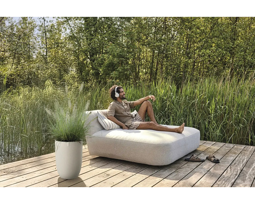 Man ontspant op een tuinligbed met een hoofdtelefoon, naast hem een plantenbak met siergras op een houten terras.