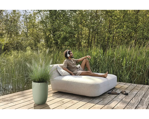 Man ontspant met een koptelefoon op een tuinbank op een houten terras naast een plantenbak met siergras.