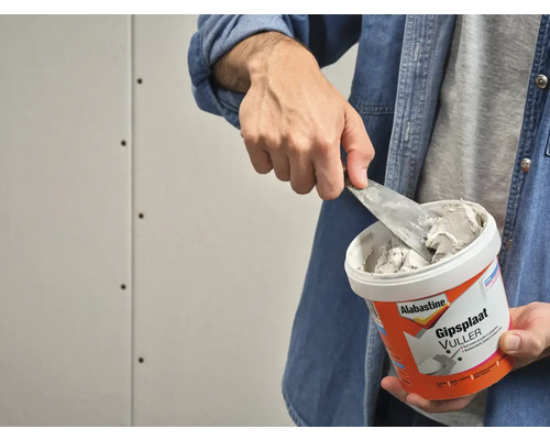 Een man neemt met een plamuurmes gipsplaatvuller uit een bak.