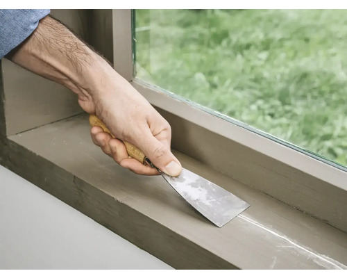 Een persoon gebruikt een plamuurmes om verf van een raamkozijn af te schrapen.