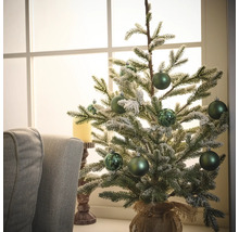 Decoratieve kerstboom met groene ballen en jutezak op een tafel voor een raam.